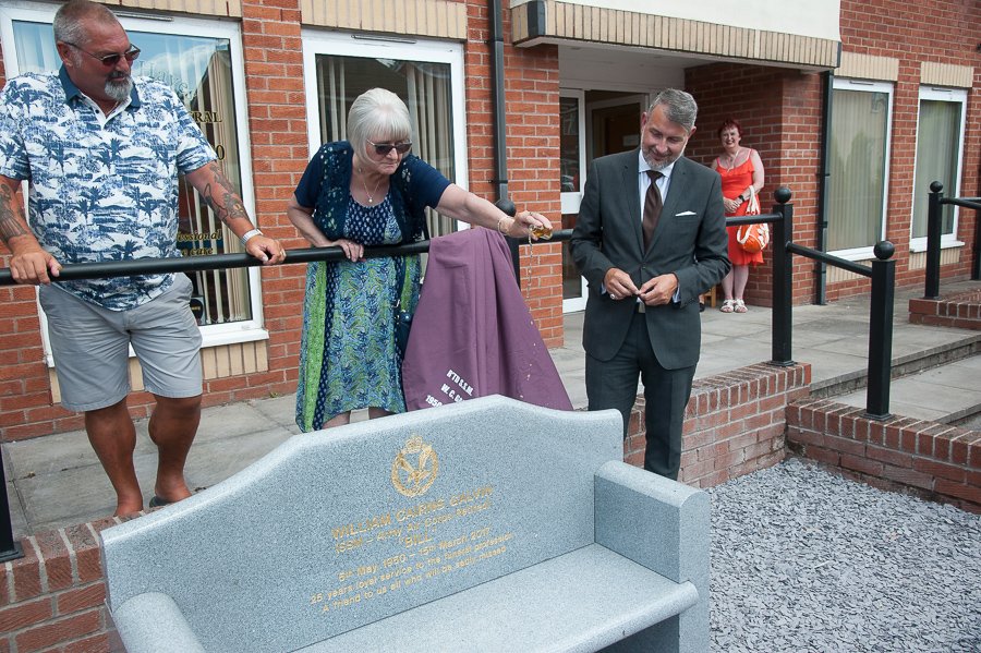 Gladys Galvin pours tot of whisky over bench erected in memory of her late husband Bill, funeral director for many years, watched by friend, Derek Barnbrook (left) and Glen Speak, deputy general manager of Halliwell funeral service.