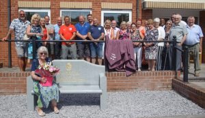 Gladys Galvin sitting on bench in memory of her late husband Bill after being presented with bouquet of flowers by his former colleagues. Looking on are villagers and Halliwell staff, including general manager Amanda Woodward (second from left) who attended unveiling of seat in Heath Hayes.